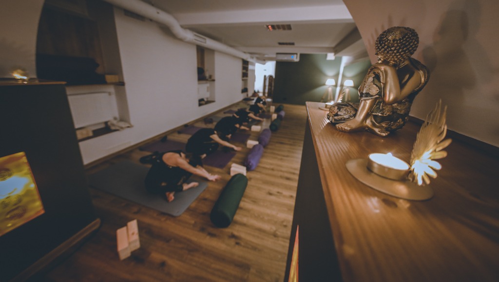 Grupa osób wykonuje pozycję dziecka na matach w studiu z drewnianą podłogą; w tle posąg Buddy i zapalona świeca na drewnianym blacie.