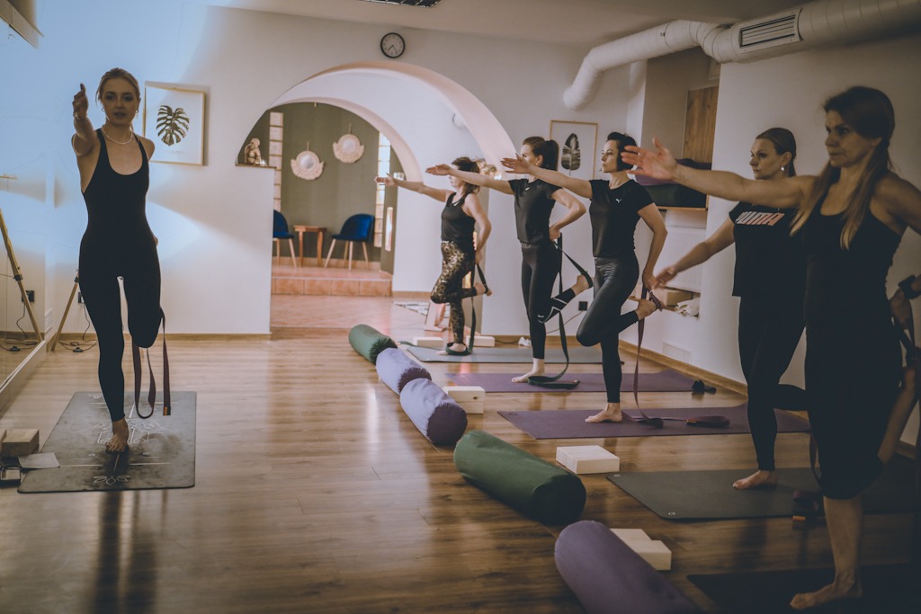 Grupa kobiet ćwiczy równowagę w studio, używając pasków do jogi, z widocznymi wałkami i kostkami do jogi na drewnianej podłodze w tle łukowatego przejścia i dekoracji ściennych.