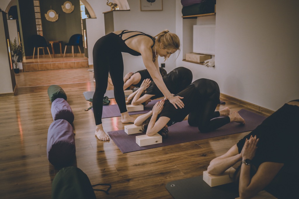 Instruktorka w czarnym stroju poprawia pozycję ćwiczącej osobie, która opiera czoło na drewnianym klocku podczas zajęć w jasnym studio z drewnianą podłogą. W tle widoczne inne osoby w podobnych...