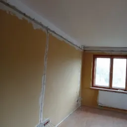 Wnętrze pokoju w Mszanie Dolnej w trakcie prac elektrycznych: widoczne kable wystające ze ściany, przygotowane pod gniazdka, oraz listwy pod sufitem z przymocowanymi przewodami.