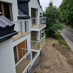 Nowoczesny budynek z balkonami wykończonymi jasnym drewnem, szklane balustrady, widok z góry na teren budowy i okoliczną drogę z zielenią.
