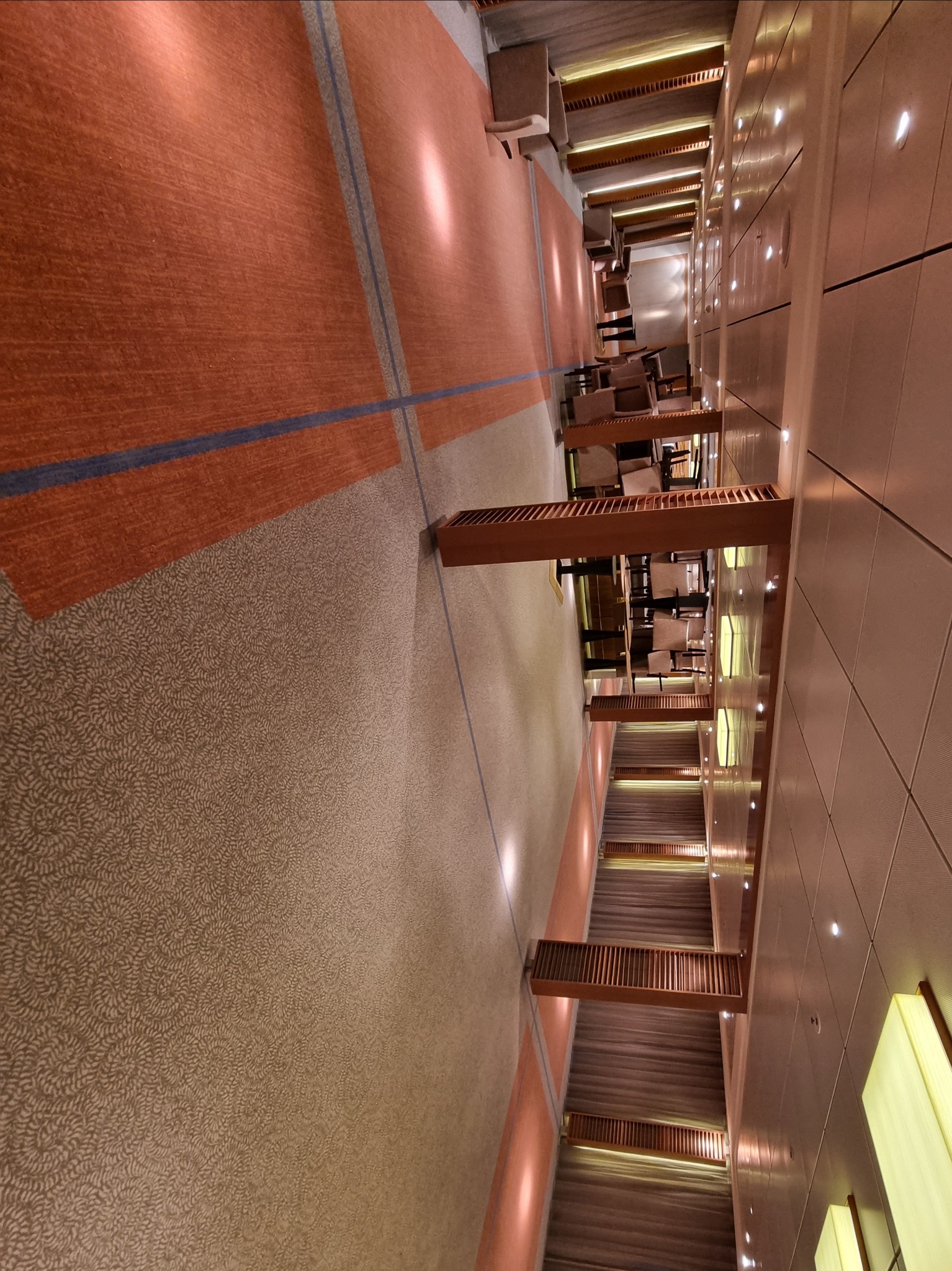 Długi korytarz hotelowy z wykładziną dywanową w odcieniach brązu i beżu, podświetlony punktowymi lampami w suficie i bocznych ścianach, prowadzący do sali konferencyjnej z widocznymi krzesłami...