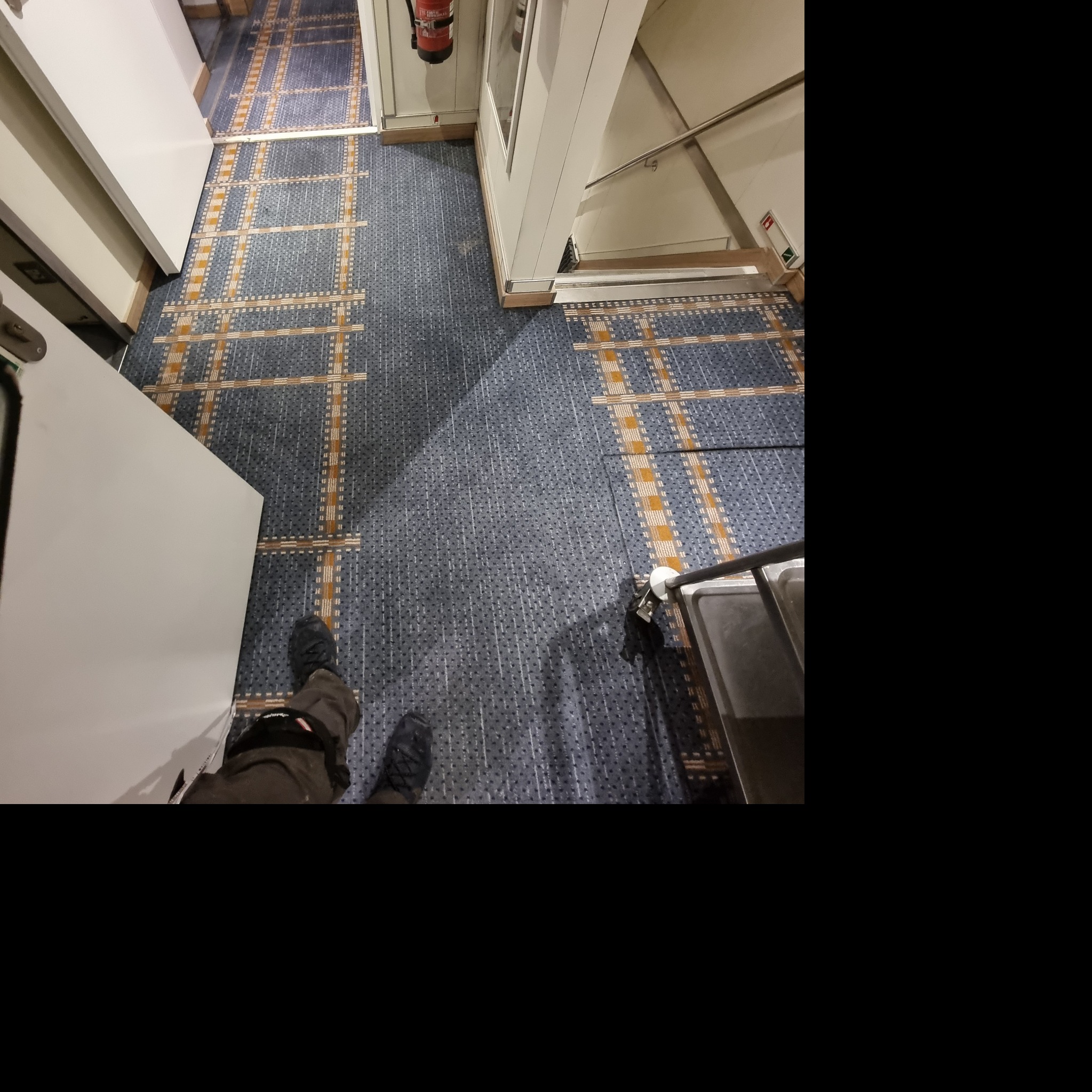 Wnętrze z nową, wzorzystą wykładziną dywanową w odcieniach szarości i beżu, widok z góry na fragment podłogi z butami i fragmentem wózka serwisowego, obok białe drzwi i ściana.