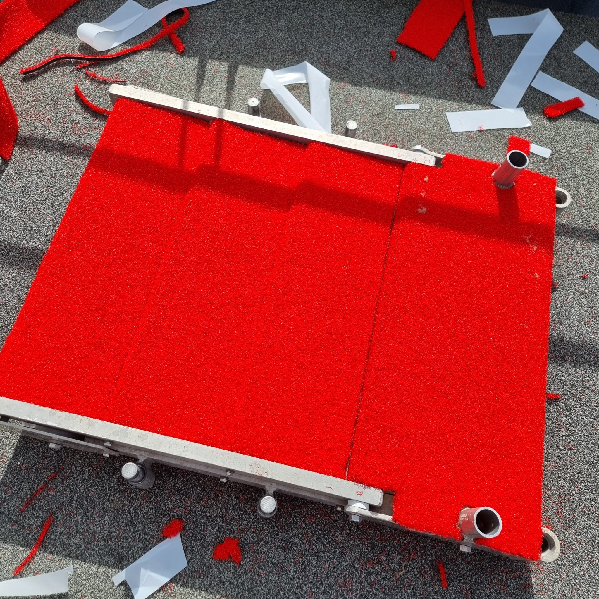 Czerwona wykładzina dywanowa z metalową ramą, fragmenty odciętej wykładziny i taśma montażowa na szarej podłodze, Gdynia.