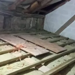 Poddasze w trakcie ocieplania: wełna mineralna między krokwiami, widoczne deski podłogowe i zwisający pomarańczowy kabel, okno dachowe w g&oacute;rnej części kadru.