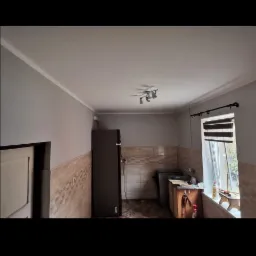 Wnętrze kuchni przed remontem, widoczne stare płytki na ścianach, ciemna lodówka i okno z roletami.