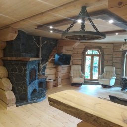 Serhii Cholavyn - Wnętrze domu z bali z kamiennym kominkiem, drewnianym żyrandolem w kształcie koła od wozu, kremowymi fotelami i widokiem na zewnątrz przez okno.