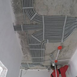 Serhii Cholavyn - Równoległe rzędy szarych, karbowanych rur instalacyjnych przymocowanych do surowego, betonowego sufitu za pomocą białych uchwytów; widoczne także przewody elektryczne i fragment osoby w czerwonej...