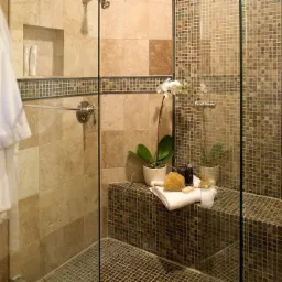 Nowoczesna kabina prysznicowa z beżowymi płytkami, mozaiką i szklaną ścianą, z rośliną w doniczce i akcesoriami do pielęgnacji na siedzisku.