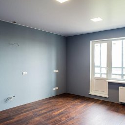 Serhii Cholavyn - Pokój po malowaniu na szaro z drewnianą podłogą, białymi drzwiami balkonowymi, grzejnikiem i wystającymi kablami elektrycznymi.