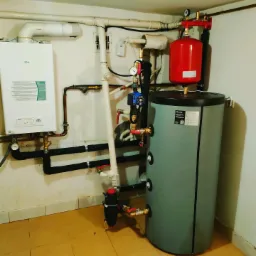 Instalacja centralnego ogrzewania w kotłowni, widoczny piec gazowy, zbiornik akumulacyjny, naczynie wzbiorcze, rury z izolacją, manometry i zawory.