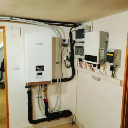 Biały, wiszący bojler Kaisai z podłączonymi rurami w izolacji, obok skrzynki elektryczne i inwertery fotowoltaiczne na ścianie w pomieszczeniu technicznym.