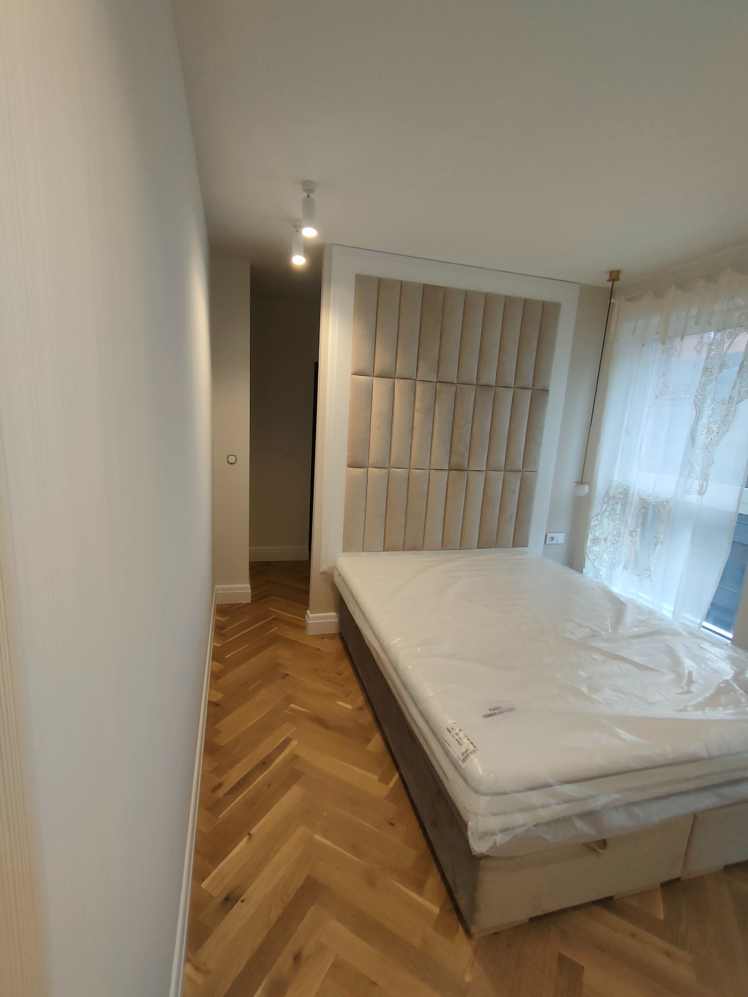 Nowoczesna sypialnia z łóżkiem z tapicerowanym wezgłowiem w pionowe pasy, drewnianą podłogą ułożoną w jodełkę i białymi ścianami. Łóżko jest jeszcze w folii ochronnej.