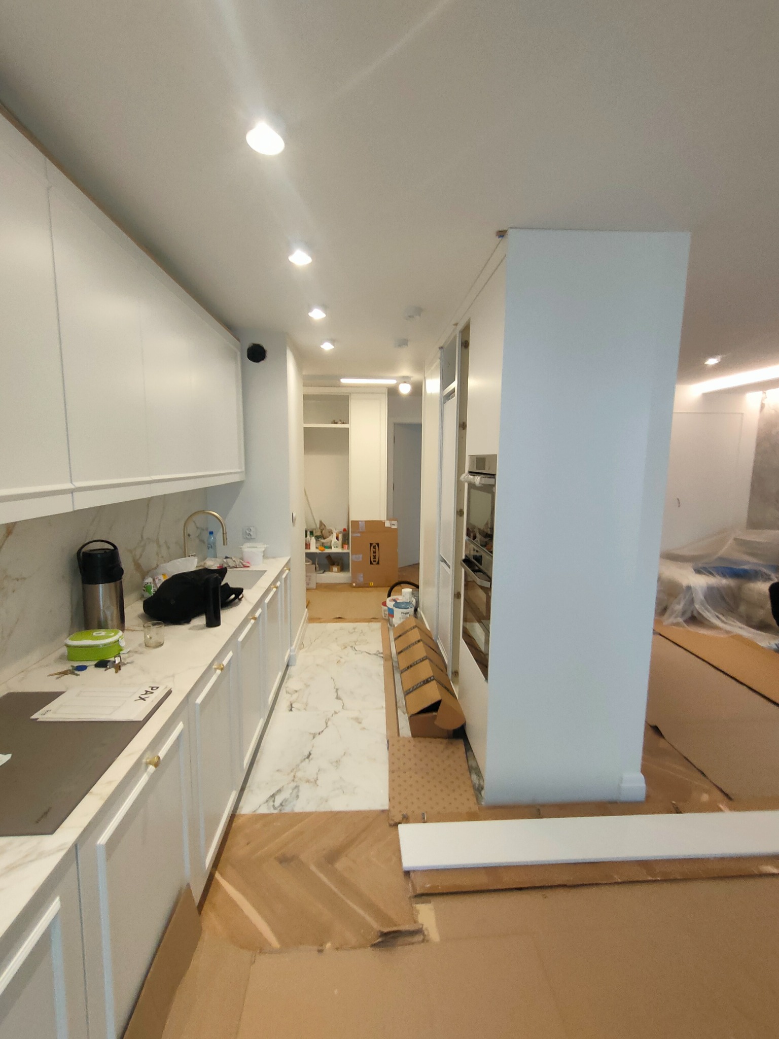 Wnętrze mieszkania w trakcie wykończenia: białe szafki kuchenne z marmurowym blatem, fragment podłogi z jasnego marmuru przechodzącego w parkiet w jodełkę, zabezpieczone kartonem podłoże, widoczne...
