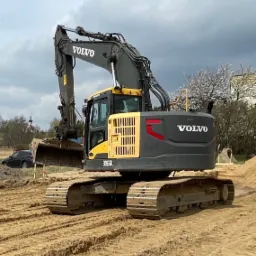 Żółto-szara koparka Volvo EC235DL na placu budowy z widocznymi śladami gąsienic w piasku i kopułą kościoła w tle.