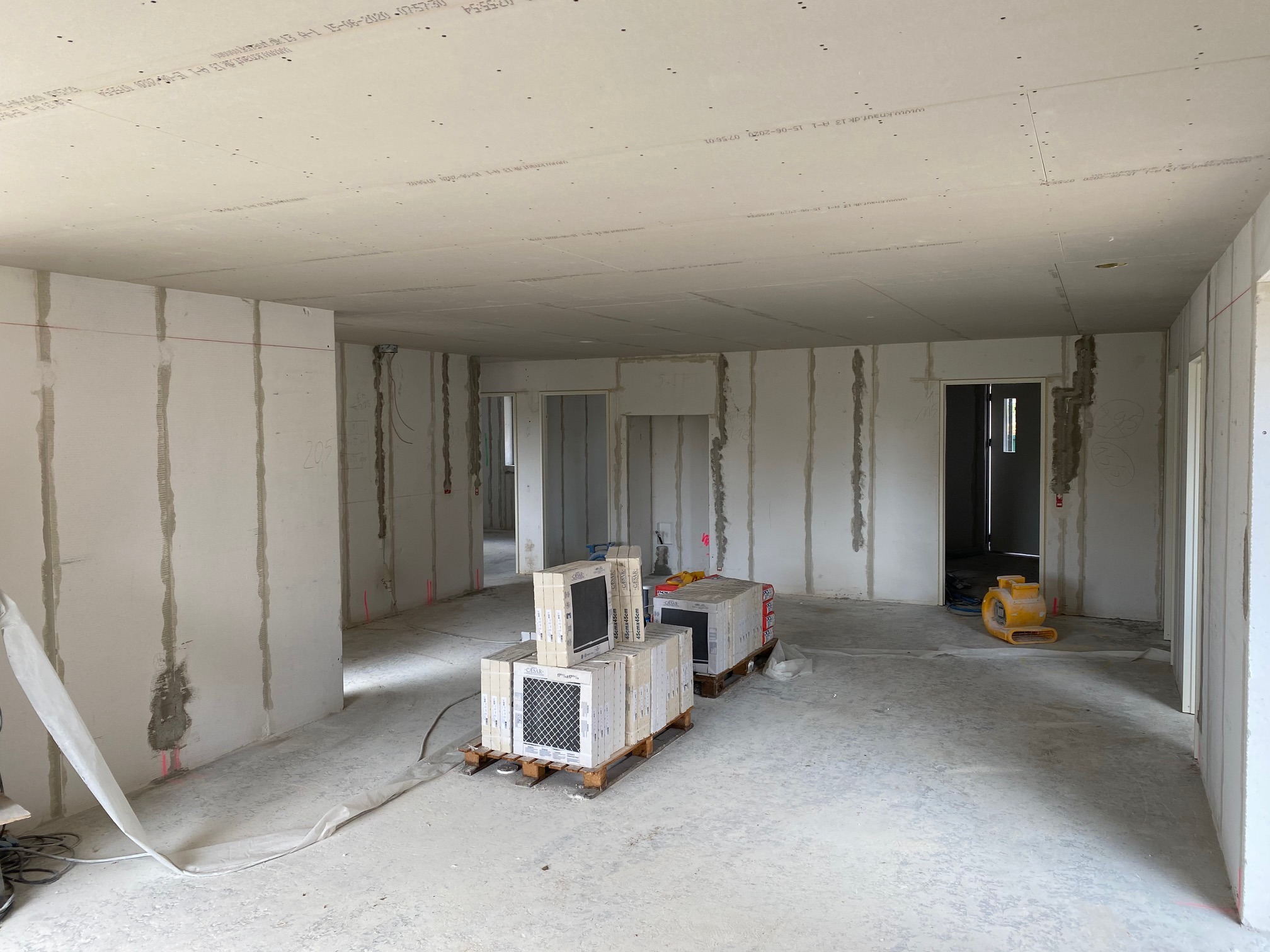 Wnętrze w trakcie prac wykończeniowych: surowe ściany z widocznymi łączeniami płyt, palety z zapakowanymi płytkami podłogowymi i nagrzewnica w tle.