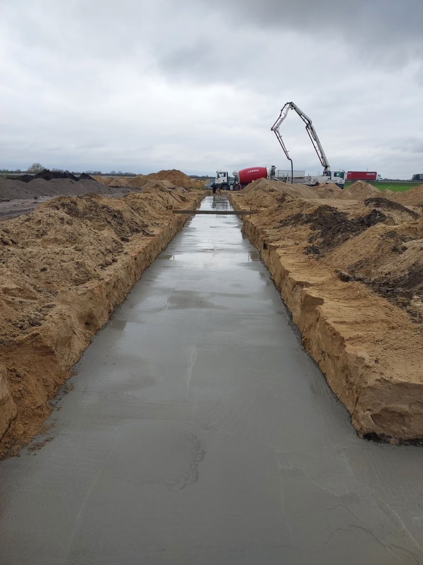 Wykop wypełniony świeżym, mokrym betonem, z widocznymi śladami szalunku po bokach, w tle betonomieszarki i pompa do betonu na tle pochmurnego nieba.