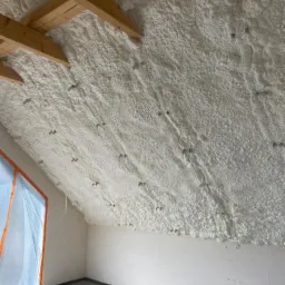 Poddasze z widoczną, grubą warstwą białej pianki izolacyjnej, aplikowaną między krokwiami drewnianymi, z widocznymi metalowymi elementami mocującymi, okno zabezpieczone folią malarską, surowy beton...