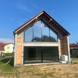 Surowy stan domu jednorodzinnego z dużymi oknami i widoczną konstrukcją dachu na tle błękitnego nieba, widoczne pustaki i elementy ocieplenia na ścianach.