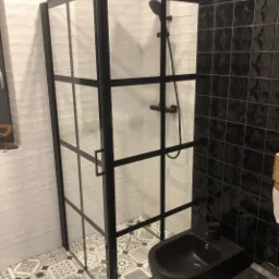 Nowoczesna łazienka z czarną kabiną prysznicową z ramami, białą cegiełkową ścianą, czarnymi kwadratowymi płytkami oraz wzorzystą podłogą w stylu patchwork.