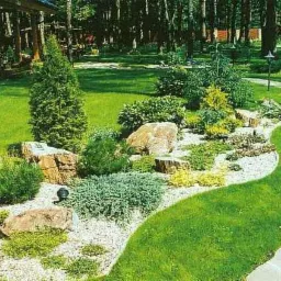 Aranżacja ogrodu z wysypanym białym żwirem, dużymi kamieniami i różnorodnymi roślinami iglastymi oraz krzewami, widoczna ścieżka z kamienia.