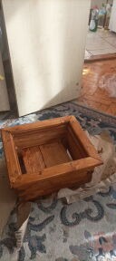 Drewniana, kwadratowa donica w odcieniach brązu, umieszczona na dywanie w pomieszczeniu. Widoczne ślady farby lub bejcy.