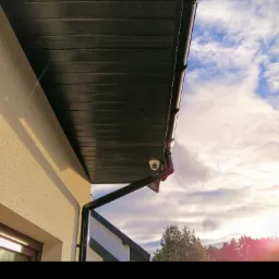 Kamera monitoringu zamontowana pod okapem budynku z widocznymi rynnami, na tle błękitnego nieba z chmurami.