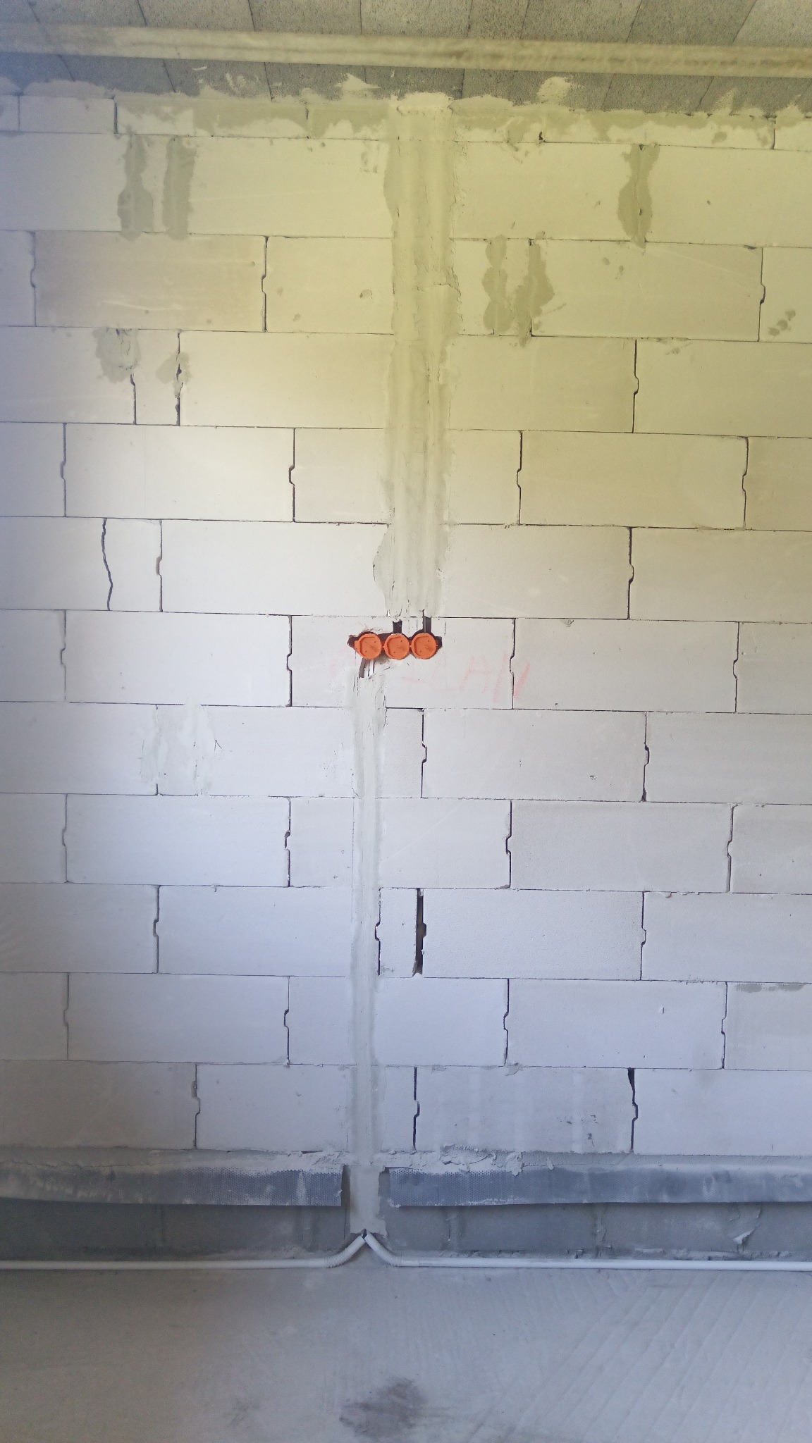 Ściana z betonu komórkowego z wyżłobionymi bruzdami pionowymi, w których umieszczono rury instalacyjne i trzy pomarańczowe puszki elektryczne podtynkowane gipsem.
