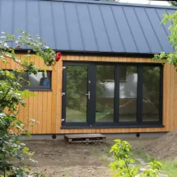 Nowoczesny budynek z drewnianą elewacją, antracytowym dachem i dużymi, czarnymi drzwiami przesuwnymi, widoczne róże w ogrodzie.