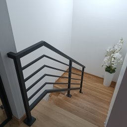 Ur-Stal Waldemar Urbaniak - Nowoczesne schody drewniane z czarną, metalową balustradą o minimalistycznym designie, doświetlone naturalnym światłem, z dekoracyjnym białym kwiatem w doniczce.