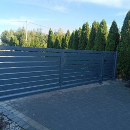 Ur-Stal Waldemar Urbaniak - Nowoczesna, przesuwna brama wjazdowa z poziomymi panelami, pomalowana na grafitowy kolor, na tle rzędu wysokich, zielonych tuj oraz błękitnego nieba. Brama zamontowana na kostce brukowej.