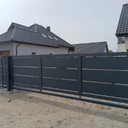 Nowoczesna, przesuwna brama wjazdowa w kolorze grafitowym z poziomymi, metalowymi akcentami, na tle domów jednorodzinnych w trakcie wykończenia.