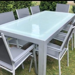 Ur-Stal Waldemar Urbaniak - Zestaw mebli ogrodowych: stół z mleczną szybą i osiem krzeseł z szarego materiału, ustawione na trawniku przed zielonym żywopłotem.