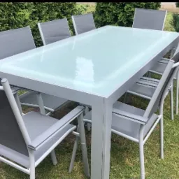 Zestaw mebli ogrodowych: stół z mleczną szybą i osiem krzeseł z szarego materiału, ustawione na trawniku przed zielonym żywopłotem.
