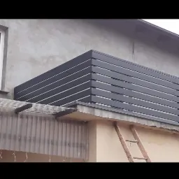 Nowoczesna, grafitowa balustrada balkonowa z poziomych profili, widoczna z dołu, z drewnianą drabiną opartą o ścianę budynku.