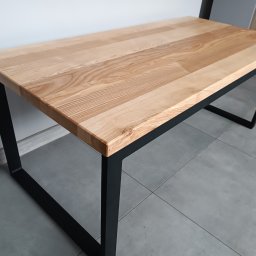 Ur-Stal Waldemar Urbaniak - Nowoczesny stół z blatem z jasnego drewna i czarnymi, metalowymi nogami o geometrycznym kształcie, ustawiony na szarych płytkach.