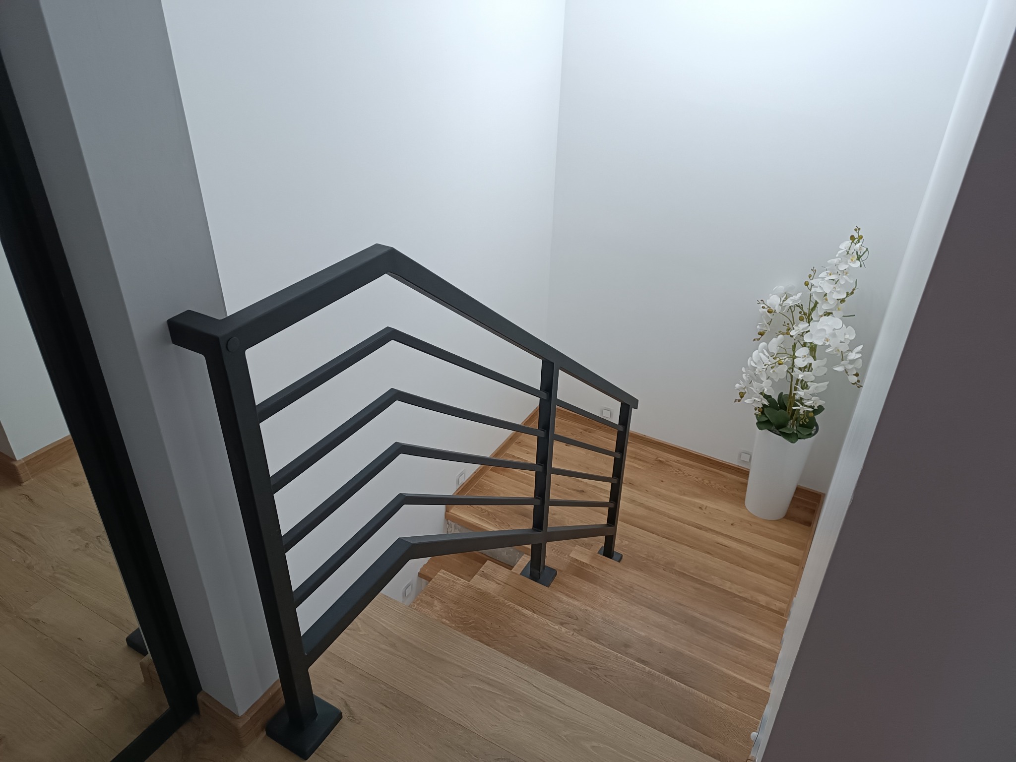 Nowoczesne schody drewniane z czarną, metalową balustradą o minimalistycznym designie, doświetlone naturalnym światłem, z dekoracyjnym białym kwiatem w doniczce.