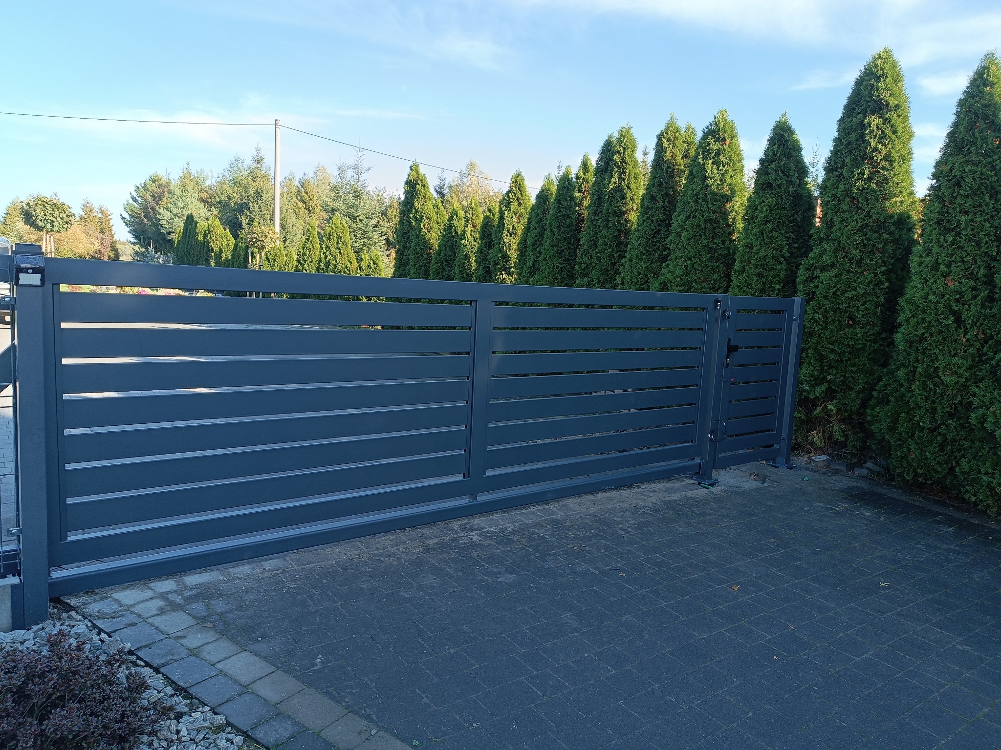 Nowoczesna, przesuwna brama wjazdowa z poziomymi panelami, pomalowana na grafitowy kolor, na tle rzędu wysokich, zielonych tuj oraz błękitnego nieba. Brama zamontowana na kostce brukowej.