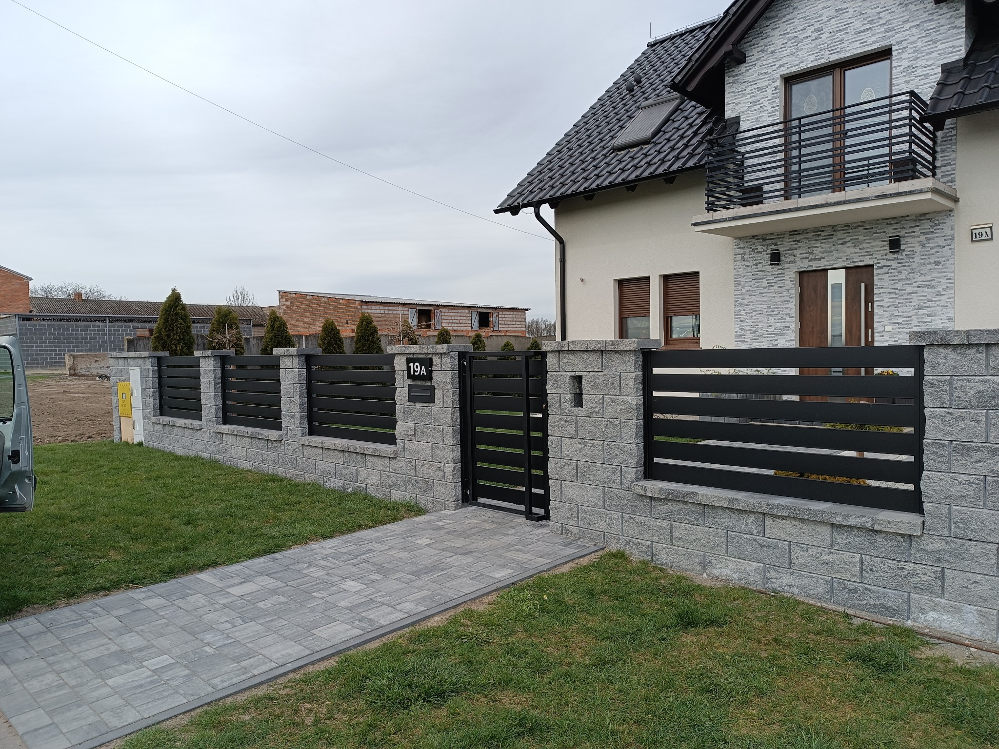 Nowoczesne ogrodzenie posesji z szarej kostki i czarnych, poziomych paneli, brama wejściowa z numerem domu 19A, na tle domu z szarą elewacją i balkonem z czarną balustradą.
