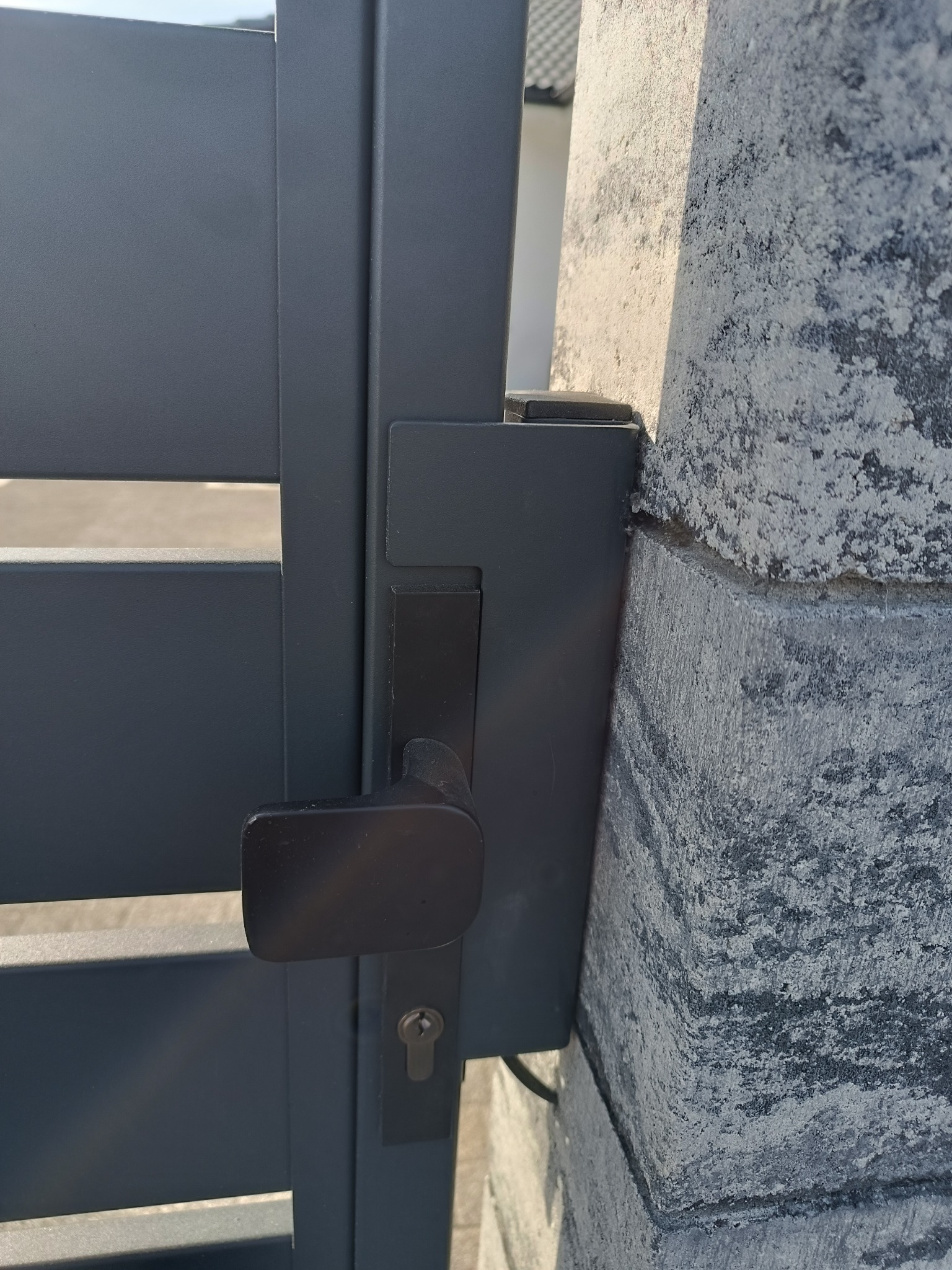 Szczegółowe zbliżenie na klamkę i zamek w nowoczesnej, metalowej bramie wjazdowej, zamontowanej na kamiennym słupku z wyraźną fakturą.