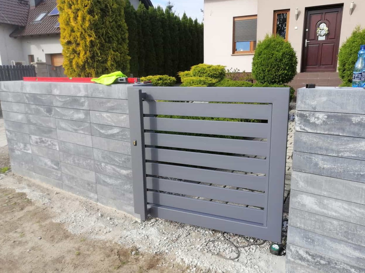 Szara, pozioma brama wjazdowa z prefabrykowanych betonowych elementów, z widoczną poziomicą i ubraniem roboczym na murku oraz domem w tle.