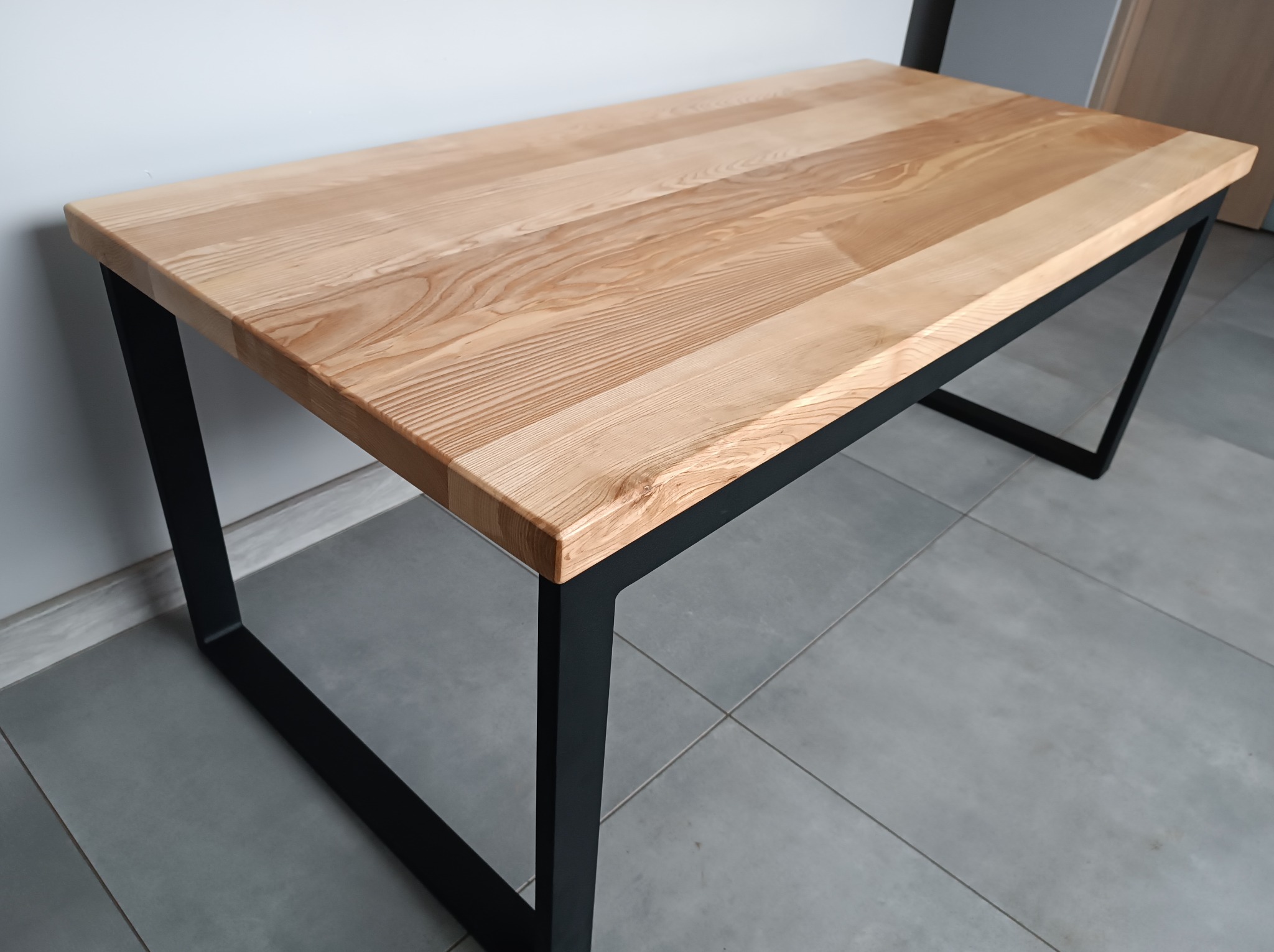 Nowoczesny stół z blatem z jasnego drewna i czarnymi, metalowymi nogami o geometrycznym kształcie, ustawiony na szarych płytkach.