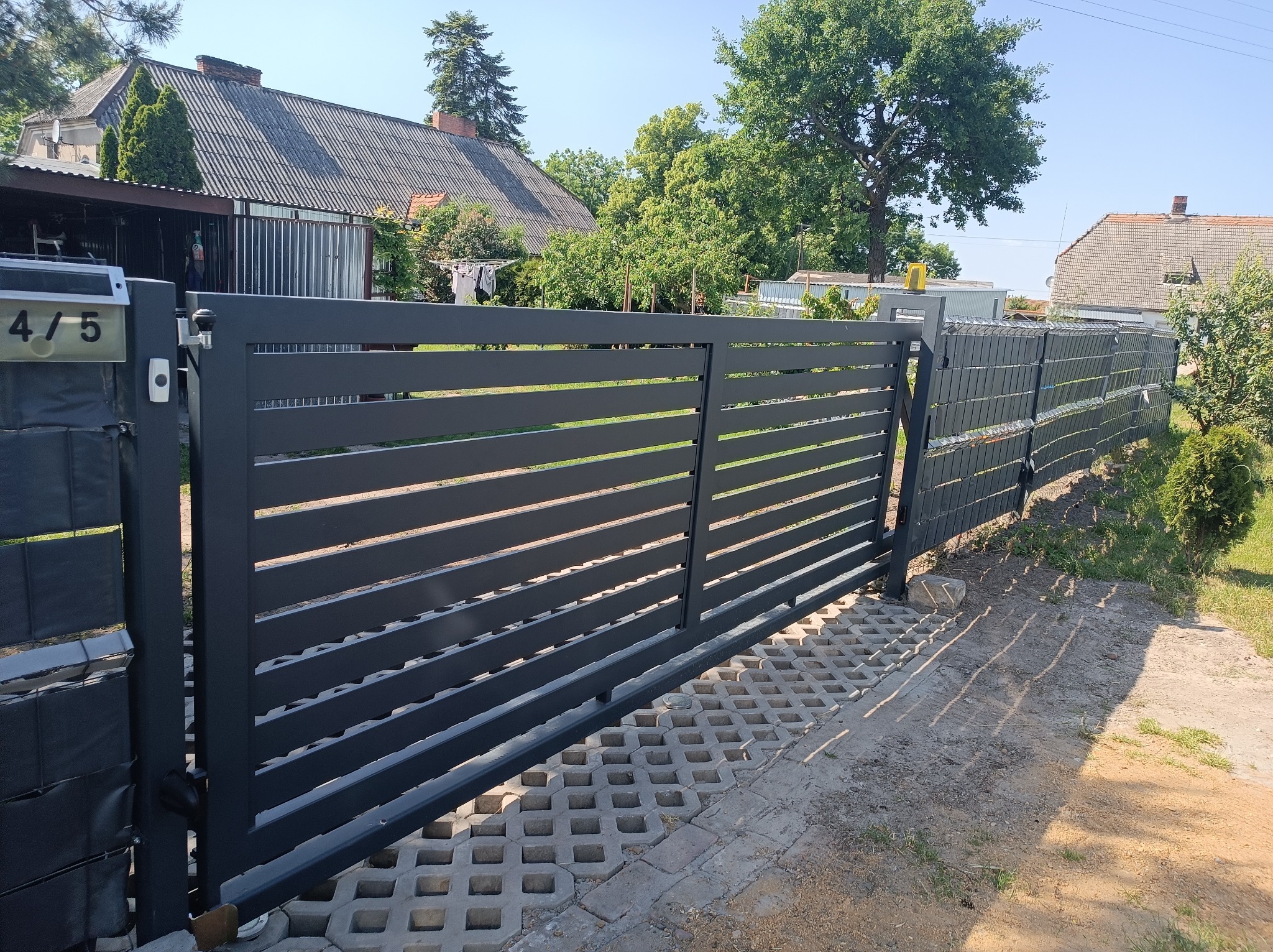 Nowoczesna, pozioma brama wjazdowa w kolorze grafitowym, z widocznym numerem posesji 4/5, osadzona na betonowych ażurowych płytach, z fragmentem ogrodzenia panelowego w tle i zielenią ogrodu.