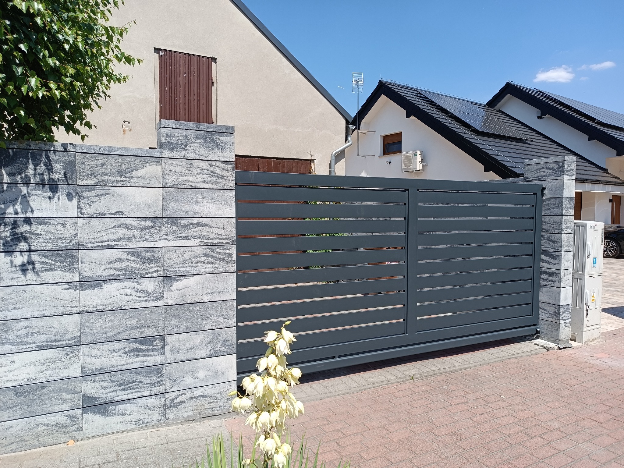 Nowoczesna, przesuwna brama wjazdowa w kolorze grafitowym, z poziomymi panelami, osadzona w murze z szarej, ciosanej kostki. Na pierwszym planie kwitnąca juka, w tle dom z panelami słonecznymi...