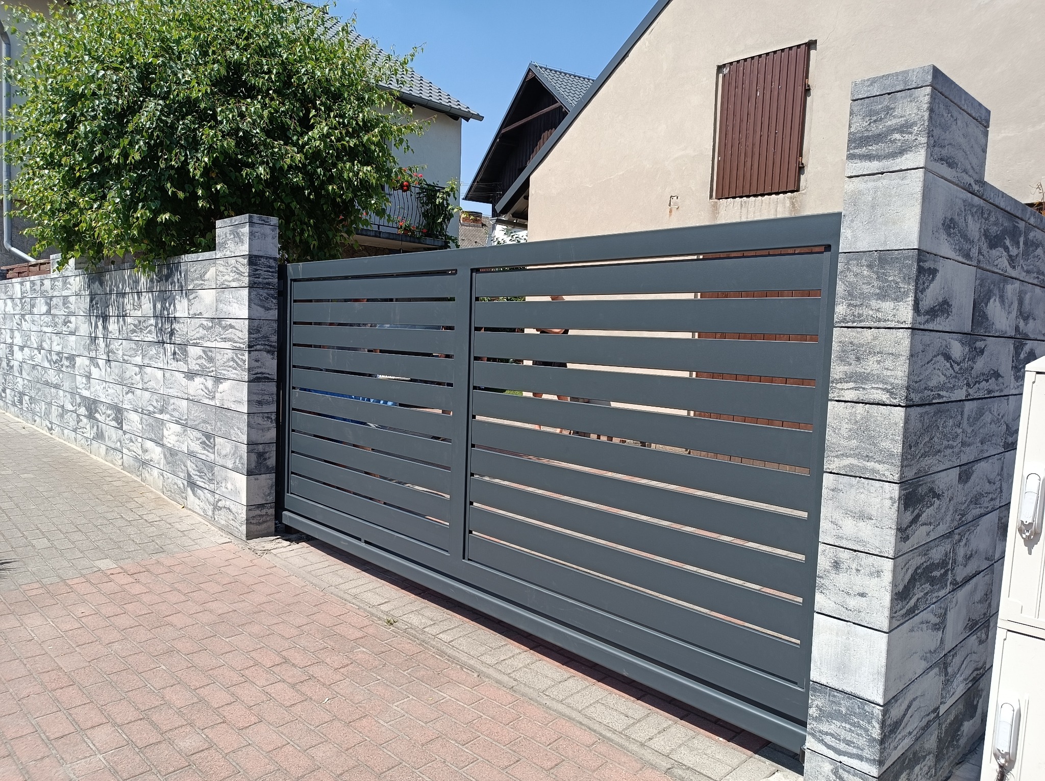 Nowoczesna, przesuwna brama wjazdowa w kolorze grafitowym z poziomymi panelami, zamontowana pomiędzy słupkami z szarej kostki imitującej kamień, na tle brukowanej drogi i zabudowań mieszkalnych.