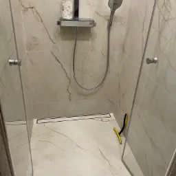 Nowoczesna kabina prysznicowa z beżowymi płytkami imitującymi marmur, odpływ liniowy, szklane drzwi i ż&oacute;łta ściągaczka do wody.