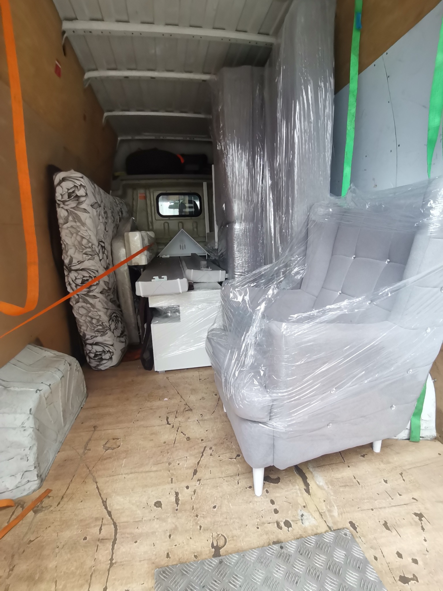 Wnętrze furgonetki załadowanej meblami owiniętymi folią stretch, w tym szary fotel z kryształkami, materac w kwiaty, białe szafki i zabezpieczające pasy transportowe.