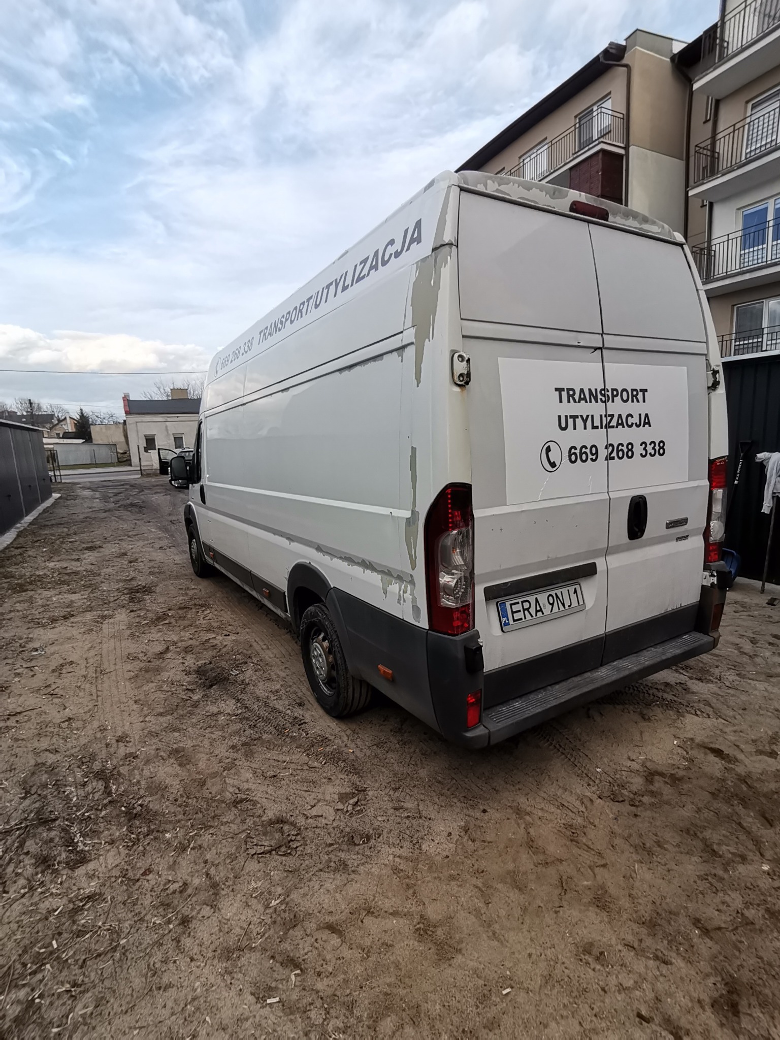 Biały van z napisem 'Transport Utylizacja' i numerem telefonu na tylnych drzwiach, zaparkowany na błotnistym terenie przed budynkiem mieszkalnym.