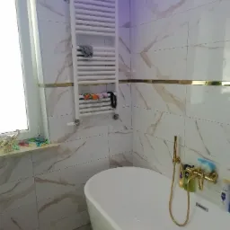 Wykończona łazienka z wanną wolnostojącą, grzejnikiem drabinkowym z ręcznikami, armaturą w kolorze złotym i białymi płytkami imitującymi marmur z złotymi paskami.