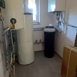 W kotłowni widoczny biały zasobnik ciepłej wody użytkowej z orurowaniem, obok biały zbiornik hydroforowy owinięty folią, czarny zmiękczacz wody, wiszący na ścianie piec gazowy kondensacyjny...